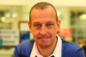 Professional headshot of man in business casual attire smiling at camera in office setting